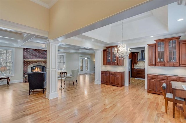 kitchen featuring ornate columns, light wood-type flooring, wall chimney range hood, a notable chandelier, and tasteful backsplash