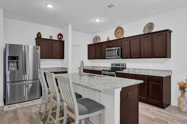 kitchen featuring light hardwood / wood-style floors, stainless steel appliances, an island with sink, dark brown cabinetry, and sink