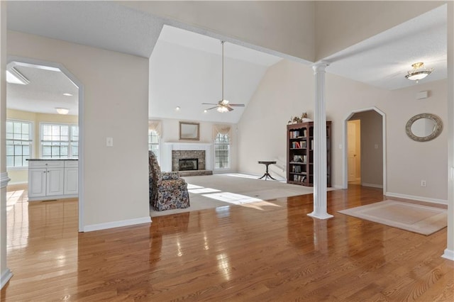 unfurnished living room featuring ceiling fan, arched walkways, a stone fireplace, light wood finished floors, and decorative columns