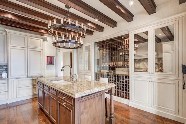 kitchen with a center island with sink, dark wood-type flooring, light stone counters, beam ceiling, and sink