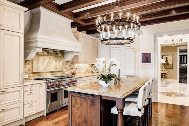 kitchen featuring an island with sink, premium range hood, light stone countertops, double oven range, and beam ceiling