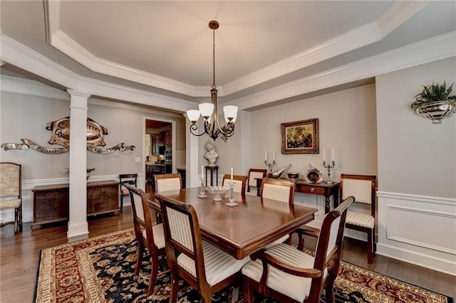 dining space featuring a wainscoted wall, wood finished floors, a tray ceiling, crown molding, and ornate columns