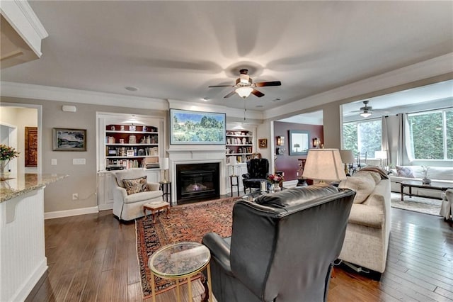 living area with dark wood-style flooring, baseboards, built in features, a glass covered fireplace, and crown molding