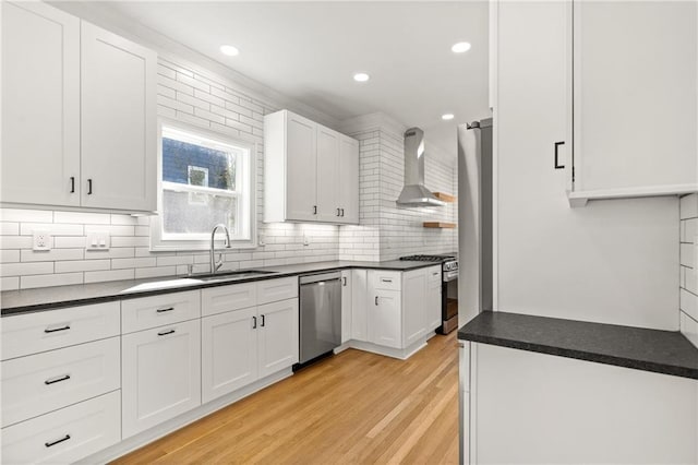 kitchen with light wood-style floors, dark countertops, stainless steel appliances, wall chimney range hood, and a sink