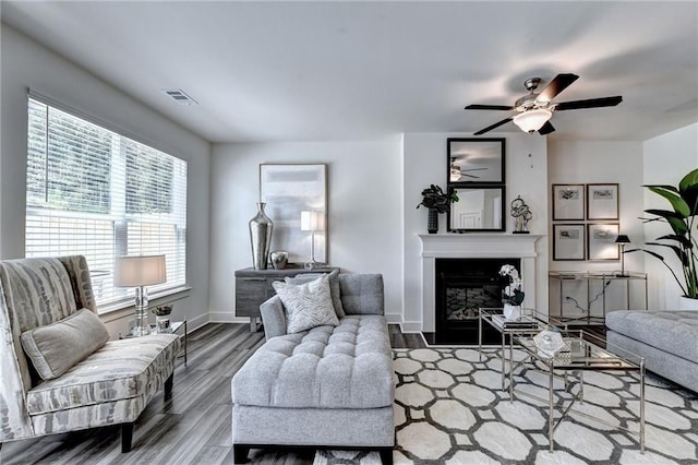 living area featuring visible vents, a glass covered fireplace, ceiling fan, wood finished floors, and baseboards