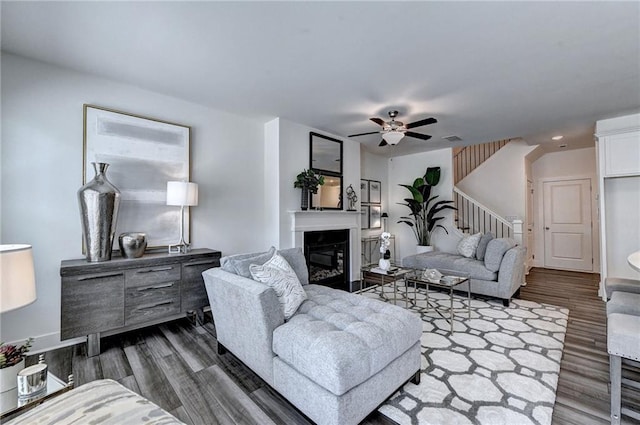 living area featuring ceiling fan, dark wood-type flooring, visible vents, stairway, and a glass covered fireplace