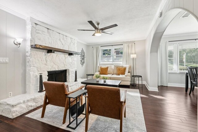 living room featuring ceiling fan, a textured ceiling, ornamental molding, dark hardwood / wood-style floors, and a stone fireplace