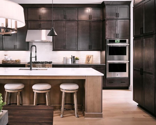 kitchen with double oven, a kitchen bar, light hardwood / wood-style floors, a center island with sink, and decorative light fixtures