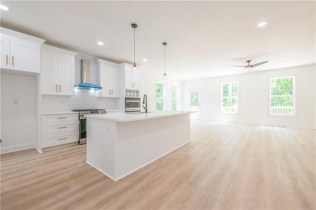 kitchen featuring white cabinetry, wall chimney exhaust hood, an island with sink, stainless steel range, and pendant lighting