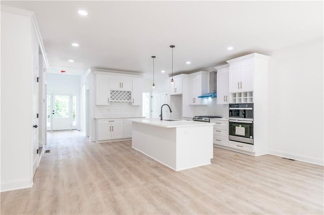 kitchen with a center island with sink, double oven, wall chimney exhaust hood, decorative backsplash, and decorative light fixtures