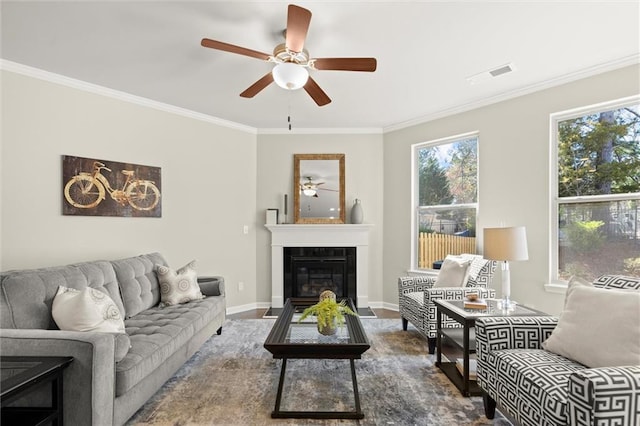 living room featuring a ceiling fan, visible vents, baseboards, ornamental molding, and a glass covered fireplace