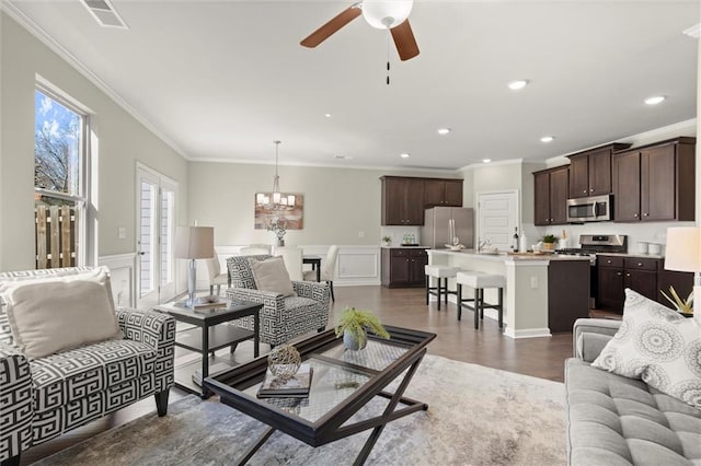 living room with dark wood-style flooring, a wainscoted wall, crown molding, visible vents, and ceiling fan with notable chandelier