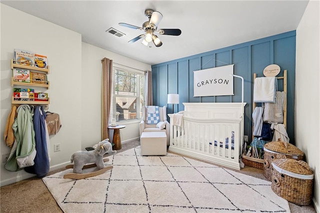 bedroom featuring a ceiling fan, carpet, visible vents, a crib, and a decorative wall