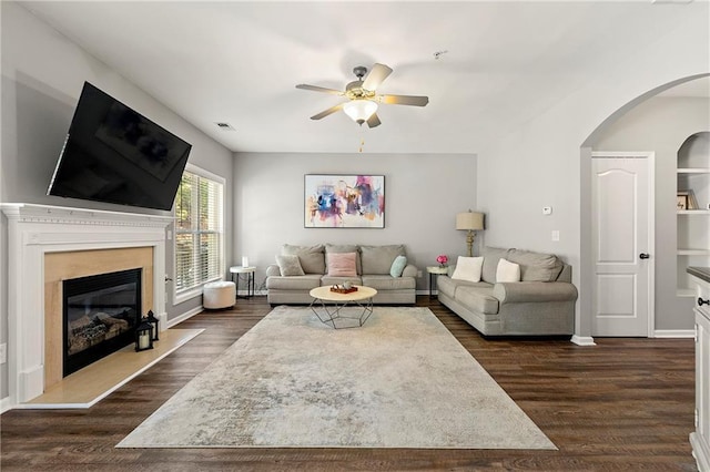 living room featuring a glass covered fireplace, dark wood-type flooring, a ceiling fan, and arched walkways