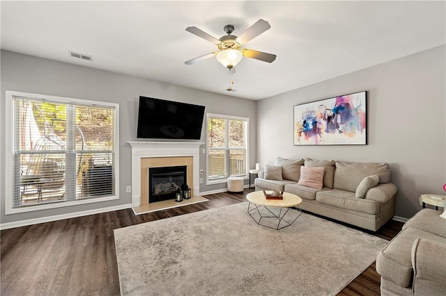 living area with visible vents, a ceiling fan, a glass covered fireplace, baseboards, and dark wood-style flooring