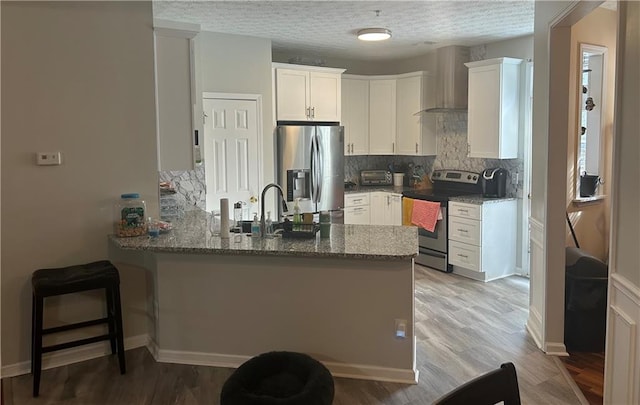 kitchen with stone counters, a peninsula, white cabinets, appliances with stainless steel finishes, and wall chimney exhaust hood