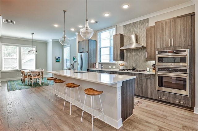 kitchen featuring visible vents, a center island with sink, a sink, stainless steel appliances, and wall chimney range hood