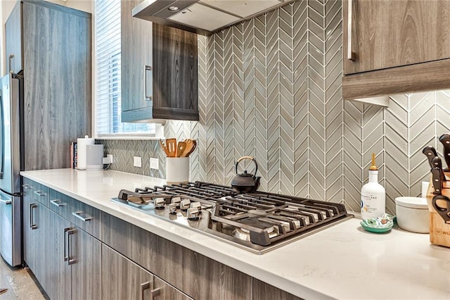 kitchen featuring backsplash, a healthy amount of sunlight, wall chimney range hood, light countertops, and stainless steel appliances