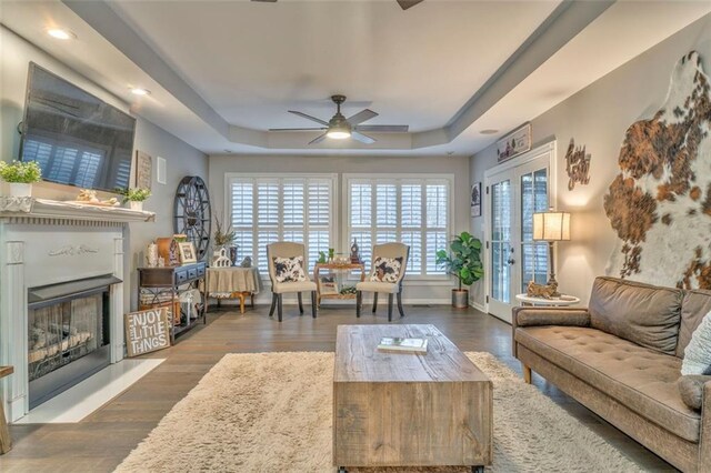 living area with a fireplace with raised hearth, baseboards, a tray ceiling, wood finished floors, and a ceiling fan