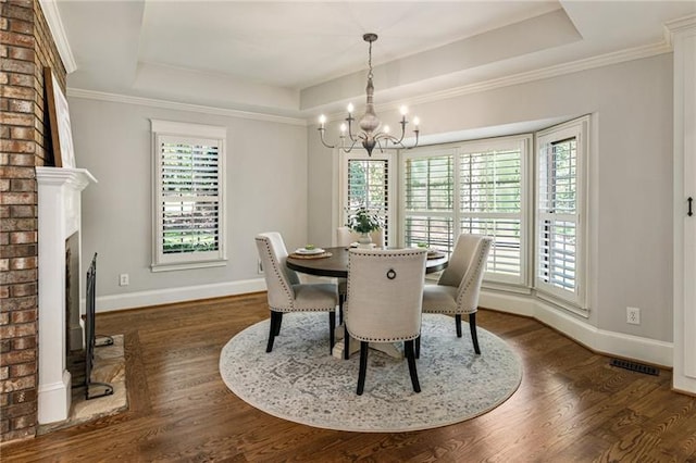 dining space featuring dark wood-type flooring, ornamental molding, a notable chandelier, a brick fireplace, and a tray ceiling