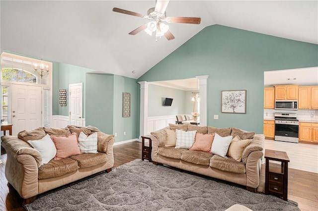 living room with high vaulted ceiling, ceiling fan with notable chandelier, a wainscoted wall, decorative columns, and wood finished floors