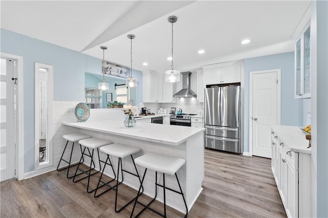 kitchen with white cabinets, a peninsula, wall chimney exhaust hood, appliances with stainless steel finishes, and a breakfast bar area
