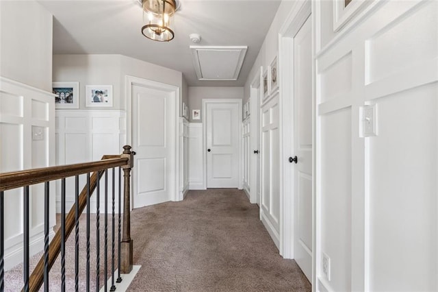 hallway featuring carpet flooring, wainscoting, a decorative wall, attic access, and an upstairs landing