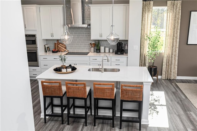 kitchen featuring wall chimney exhaust hood, a sink, wood finished floors, backsplash, and double oven