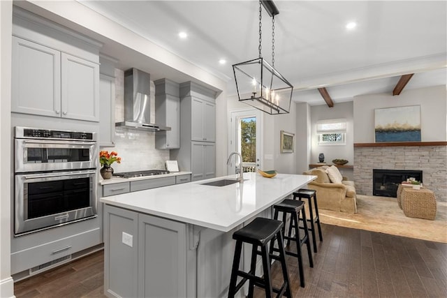 kitchen with appliances with stainless steel finishes, wall chimney exhaust hood, sink, beam ceiling, and a center island with sink