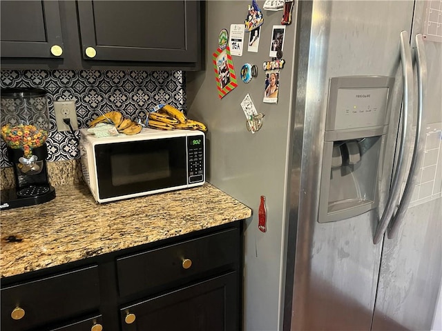 kitchen with light stone counters, decorative backsplash, dark cabinetry, and stainless steel refrigerator with ice dispenser