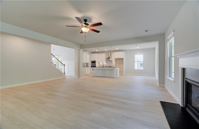 unfurnished living room with light wood-style flooring, stairway, a ceiling fan, a glass covered fireplace, and baseboards