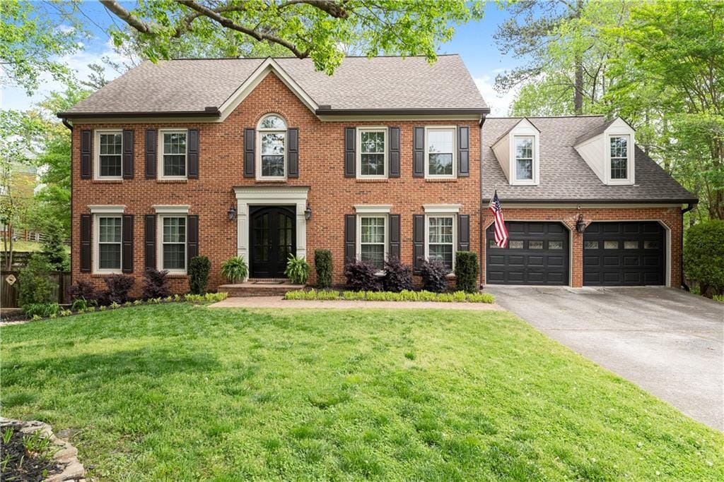 colonial house featuring a shingled roof, a front lawn, concrete driveway, an attached garage, and brick siding