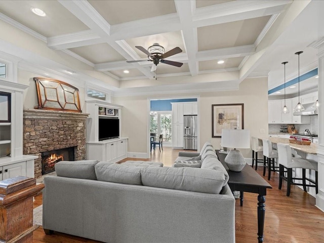 living room with a stone fireplace, hardwood / wood-style flooring, coffered ceiling, ceiling fan, and beam ceiling