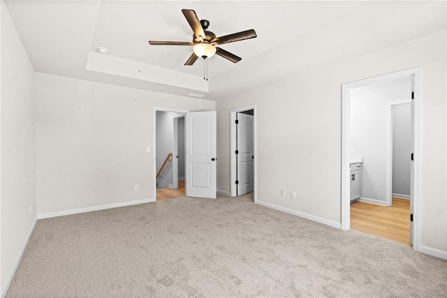 unfurnished bedroom with baseboards, ceiling fan, light colored carpet, a tray ceiling, and ensuite bath