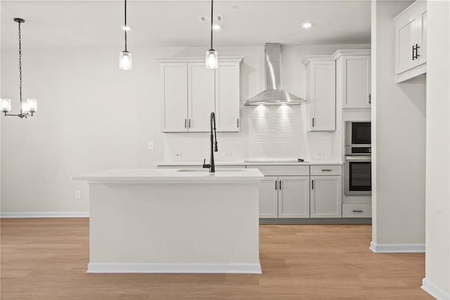 kitchen with stainless steel oven, an island with sink, wall chimney exhaust hood, and light wood finished floors