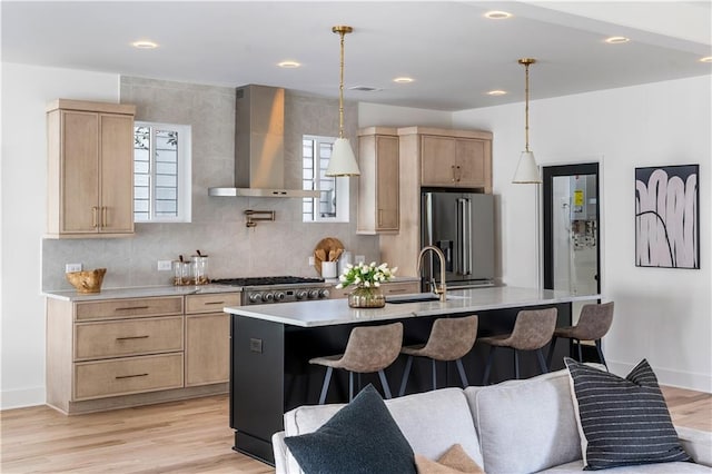 kitchen with high quality fridge, light countertops, wall chimney range hood, an island with sink, and decorative light fixtures
