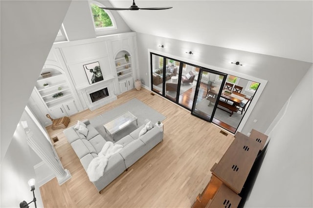 living area featuring light wood-style flooring, built in features, a fireplace with raised hearth, high vaulted ceiling, and a ceiling fan