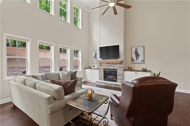 living room with ceiling fan, dark wood finished floors, a stone fireplace, baseboards, and a towering ceiling
