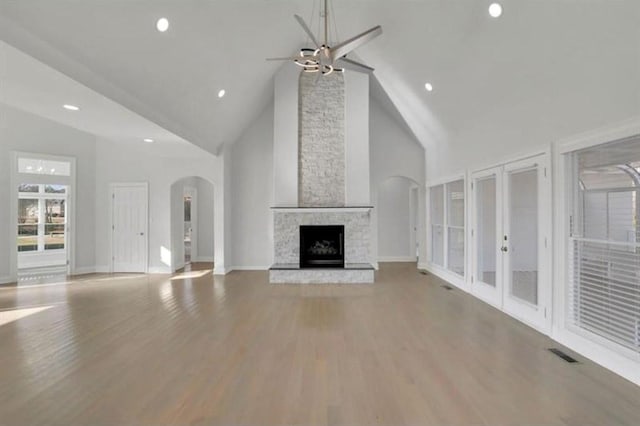 unfurnished living room featuring arched walkways, ceiling fan, a stone fireplace, wood finished floors, and high vaulted ceiling