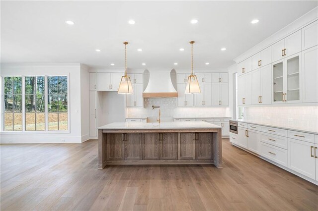 kitchen with white cabinets, stainless steel microwave, premium range hood, tasteful backsplash, and an island with sink