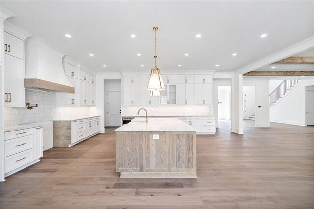 kitchen with custom range hood, a center island with sink, crown molding, and light wood-type flooring