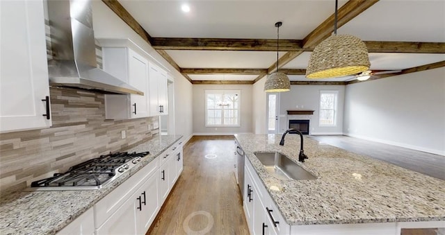 kitchen featuring wall chimney exhaust hood, sink, a center island with sink, hanging light fixtures, and stainless steel gas stovetop