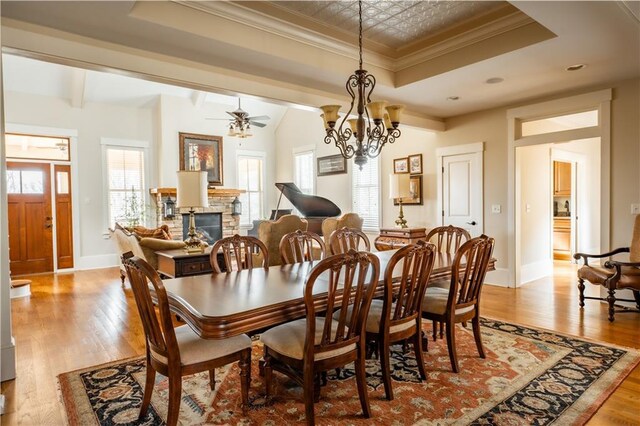 dining room with ceiling fan with notable chandelier, a fireplace, a raised ceiling, crown molding, and light wood-type flooring