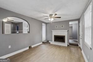 unfurnished living room with wood finished floors, a fireplace with raised hearth, a ceiling fan, and baseboards
