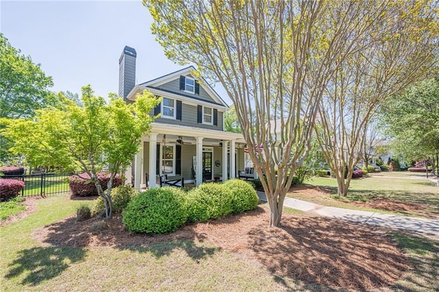 american foursquare style home featuring ceiling fan, covered porch, a chimney, fence, and a front lawn