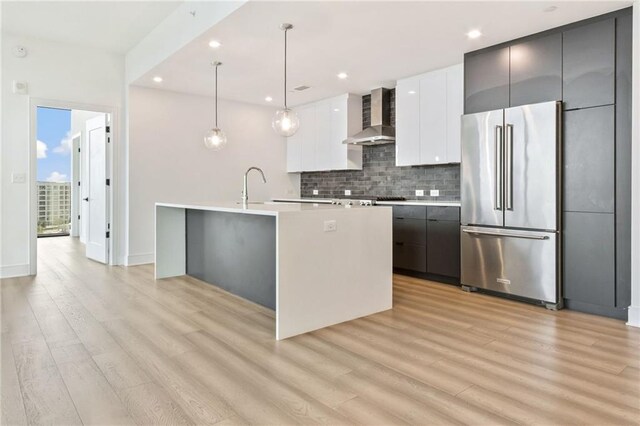 kitchen featuring white cabinets, hanging light fixtures, high end refrigerator, wall chimney exhaust hood, and a center island with sink