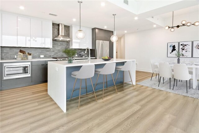 kitchen featuring tasteful backsplash, an island with sink, wall chimney range hood, light hardwood / wood-style flooring, and decorative light fixtures