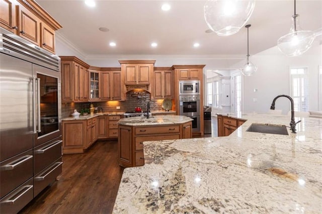 kitchen featuring built in appliances, a sink, brown cabinetry, a kitchen island with sink, and ornamental molding