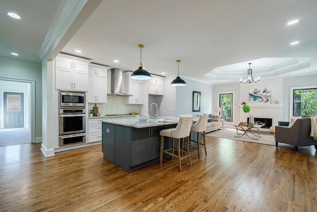 kitchen with wall chimney exhaust hood, a tray ceiling, white cabinets, stainless steel microwave, and an island with sink