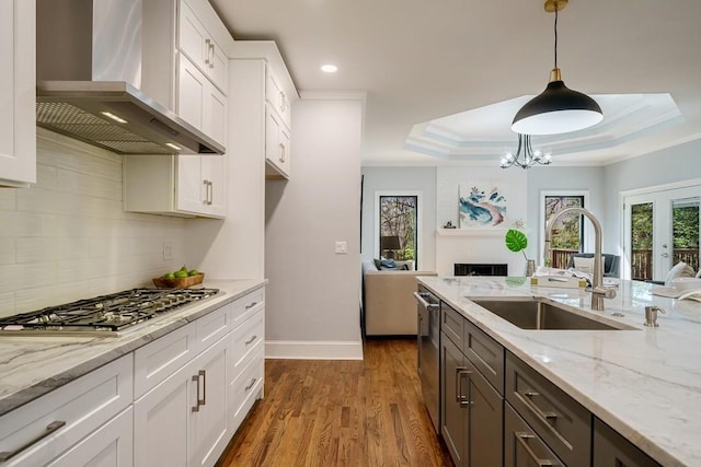 kitchen featuring appliances with stainless steel finishes, a tray ceiling, wall chimney range hood, decorative light fixtures, and white cabinetry